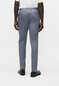 Άνδρας φοράει στενά ανοιχτό μπλε παντελόνια φόρμας, λευκό πουκάμισο μέσα, και μαύρα παπούτσια φόρμας, όρθιος γυρισμένος προς τα πίσω σε απλό φόντο.