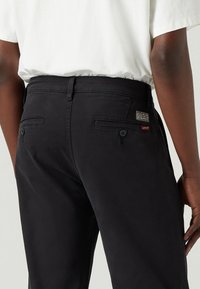 Uomo con pantaloni neri Levi's con tasche posteriori chiuse da bottoni e una camicia bianca infilata, visto dalla vita in giù, da dietro.