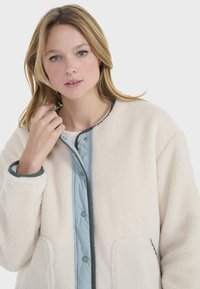 Chaqueta de forro polar en color crema con un forro azul claro, que presenta botones verdes, escote redondeado y dos bolsillos frontales. Tejido suave y texturizado.