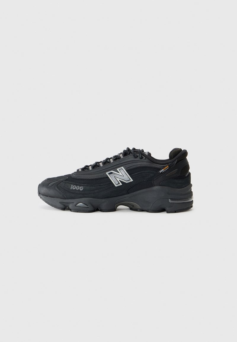 Chaussure de sport New Balance noire avec logo argenté réfléchissant, semelle épaisse et marquages "1000" et "Cordura" sur un fond clair uni.