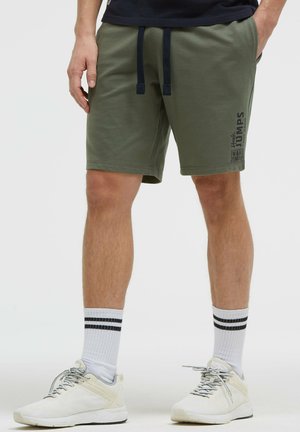 Mann trägt olivgrüne Shorts mit schwarzen Kordeln, weiße Sneakers und weiße Socken mit schwarzen Streifen, steht mit den Händen in den Taschen.