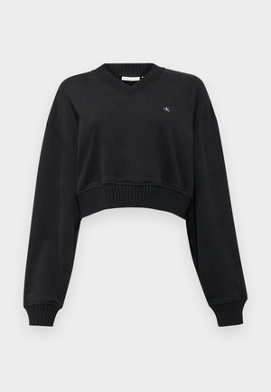 Črn skrajšan pulover s V-izrezom, dolgimi rokavi in rebrastim robom. Ima majhen vezen logotip na prsih. Mehak material.