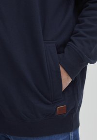 Sweatshirt bleu marine avec une poche frontale, des poignets côtelés et une étiquette en cuir détaillant la marque. Texture douce et coupe décontractée.