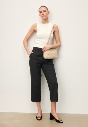 Žena stojící opřená o zeď, oblečená v bílém tílku bez rukávů, černých zkrácených kalhotách, černých sandálech s otevřenou špičkou a s béžovou prošívanou kabelkou přes rameno.