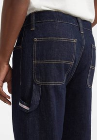Jeans de mezclilla azul oscuro con grandes bolsillos traseros cuadrados y un pequeño lazo de utilidad etiquetado "Guess Jeans" en el lado, llevados por una persona.