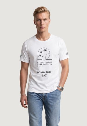 Giovane uomo con maglietta bianca "Internazionali BNL d'Italia Roma 2026" e jeans blu, in piedi con una mano in tasca.