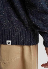 Granatowy sweter dzianinowy z kolorowymi plamkami, z prążkowanymi mankietami i dołem, z małą metką z logo. Częściowo widoczny beżowy materiał spodni.