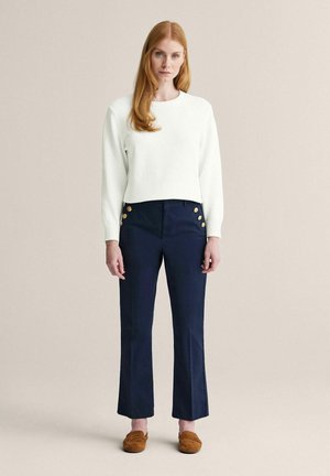 Pitkätukkainen punapää nainen, joka pukeutunut valkoiseen pitkähihaiseen neulepuseroon, laivastonsinisiin housuihin joissa on kultaiset napit, ja ruskeisiin loafereihin, seisoo yksinkertaista taustaa vasten.