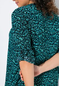 Camicia stampata leopardata turchese e nera con maniche arrotolate, dotata di un dettaglio a bottone sui polsini. Tessuto morbido con una superficie leggermente strutturata.