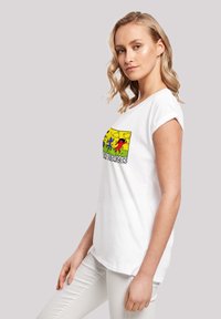 Balta medvilninė marškinėliai su trumpomis rankovėmis, suktomis į viršų, su spalvinga grafika figūrų ant geltonos fono ir tekstu "SUPER HEROJAI."