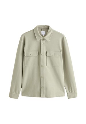 Person, der spænder den beige knaphulsknap på en langærmet jakke over en hvid skjorte.