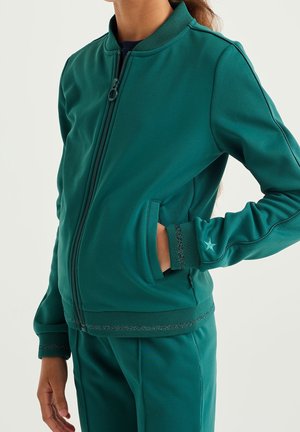 Sileästä kankaasta valmistettu teal-värinen vetoketjullinen takki, jossa on resorikaulus ja -hihansuut, sivutaskut ja koristeompeleet hihoissa.