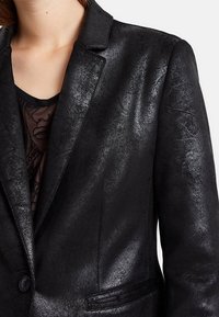 Blazer noir fabriqué en matériau synthétique texturé, avec une fermeture à un seul bouton, des revers crantés et deux poches avant.