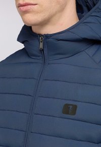 Námořnická péřová bunda s horizontálními švy, zapínáním na zip a kapucí. V oblasti hrudi má malé černé logo. Hladký povrch.