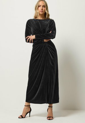 Mujer posando con vestido negro de terciopelo de manga larga con detalle fruncido en la cintura y sandalias negras de tacón alto sobre fondo neutro.