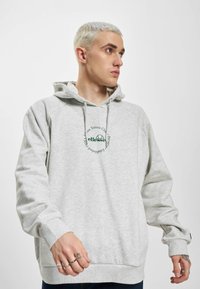 Ung mand med kort blondt hår iført lysegrå hættetrøje med grønt "ellesse" logo og tekst på brystet, kigger mod højre.