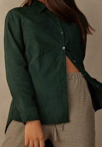 Camicetta verde scuro in tessuto morbido con due tasche sul petto e orlo curvo, abbinata a pantaloni beige a scacchi.