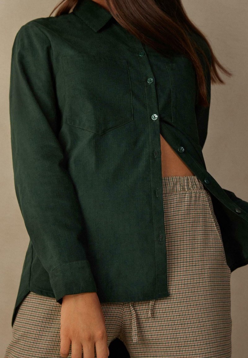 Camicetta verde scuro in tessuto morbido con due tasche sul petto e orlo curvo, abbinata a pantaloni beige a scacchi.