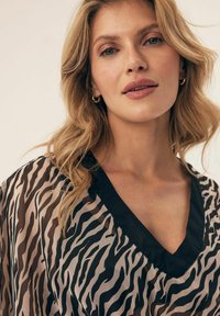 Przezroczysta bluzka w czarno-beżowy wzór zebry, zaprojektowana z dekoltem w kształcie litery V oraz czarnymi wykończeniami. Gładka faktura i zwiewny materiał.