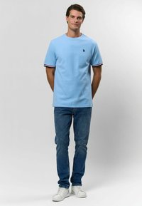 Svetlomodré bavlnené tričko s krátkymi rukávmi, so malou nášivkou na hrudi a kontrastnými pruhovanými akcentmi na manžetách.