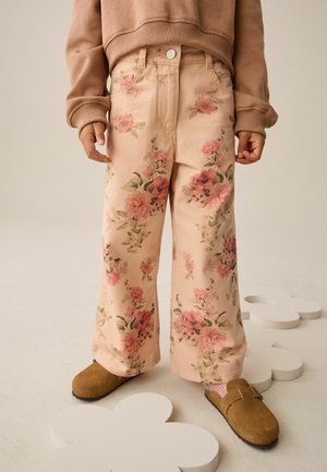Pantalones de pierna ancha con estampado floral en tela rosa suave, con flores en tonos rosa y verde. Combinados con zuecos marrones de ante y un suéter beige.
