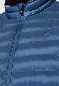 Veste matelassée en bleu foncé, avec une texture matelassée, un col haut et une fermeture éclair. Petit logo sur la poitrine en rouge, blanc et bleu.