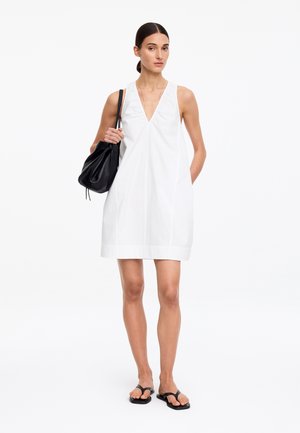 Femme aux cheveux foncés en chignon bas, vêtue d’une robe blanche sans manches à col en V, sandales noires et portant un sac à épaule noir.
