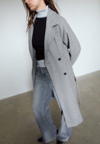 Grauer Wollmischmantel mit doppelreihigem Design, getragen über einem schwarzen Oberteil und einem hellblauen Rollkragenpullover. Kombiniert mit ausgestellt geschnittenen hellen Jeans.