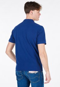 Polo blu realizzato in cotone texturizzato. Maniche corte, colletto e orlo dritto. Indossato con jeans blu, che mostrano un dettaglio della tasca posteriore.