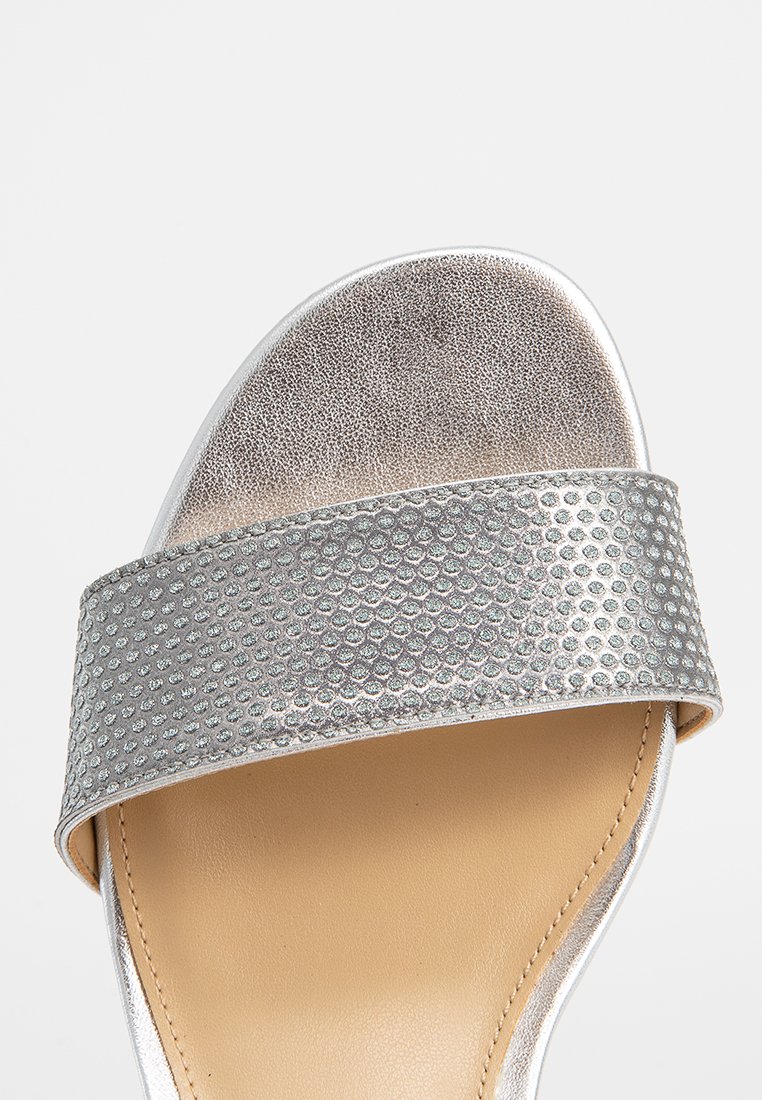 Tongs argentées avec une bride métallique texturée comportant des points surélevés, une semelle intérieure lisse et une semelle arrondie.