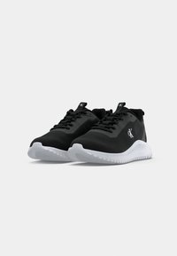 Paire de chaussures de sport noires avec semelles blanches, design à lacets et logo "CK" sur les côtés et la languette, présentées sur un fond clair uni.
