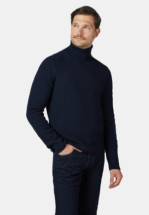 Man draagt een donkerblauwe getextureerde coltrui en donkere broek, met één hand in zijn zak, kijkt iets schuin omhoog naar de zijkant.