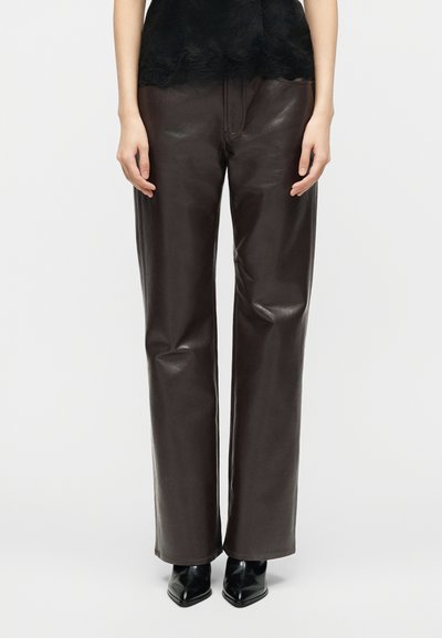 Oseba v temno rjavih usnjenih hlačah z ravnimi nogami, črni majici z čipkastim robom in črnih čevljih z ozkim sprednjim delom na belem ozadju.