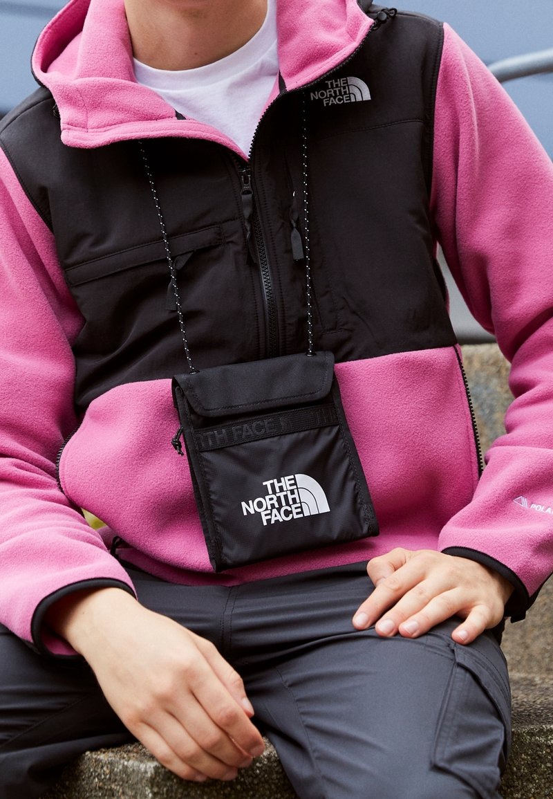 Sudadera de forro polar negra y rosa con bolsillo con cremallera, con un logo negro y un pequeño estuche negro unido por un cordón alrededor del cuello.