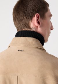 Tan suede jacket with a black ribbed collar, featuring a subtle logo on the back and clean stitching detail. Smooth texture throughout.