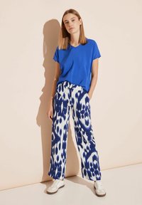 Niebieski top z krótkim rękawem w połączeniu z białymi i granatowymi spodniami w szerokie nogawki we wzory. Model nosi białe sneakersy z jasnobrązowymi akcentami.