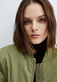 Olivgrüne Bomberjacke mit Reißverschluss, gerippten Bündchen und glatter Textur. Über einem schwarzen Rollkragenpullover getragen, mit einem Nahaufnahme des Gesichts.