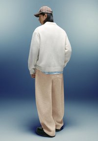 Osoba oblečená v béžových širokých nohaviciach, bielom svetri, károvanom klobúku a čiernych topánkach, stojí proti pozadiu so gradientom modrej farby a čelí bokom.