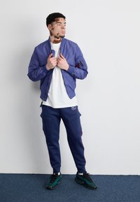 Fioletowa kurtka bombka z zamykanymi kieszeniami, biała koszulka, granatowe dresowe spodnie cargo z bocznymi paskami i zielone buty sportowe. Jednolity tle.