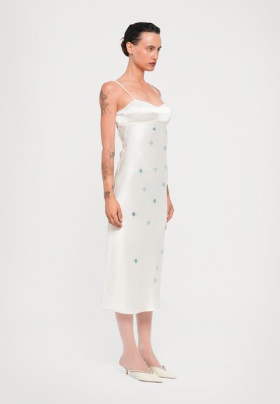 Witte satijnen slipdress met dunne spaghettibandjes, met blauwe bloemenborduursels. Zachte textuur, midi-lengte en subtiele glans. Paar witte puntige muiltjes.