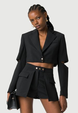 Kobieta w czarnym, krótkim blezerze z wyciętymi rękawami i dopasowanych szortach z wysokim stanem, trzymająca czarną torebkę na jednolitym tle.