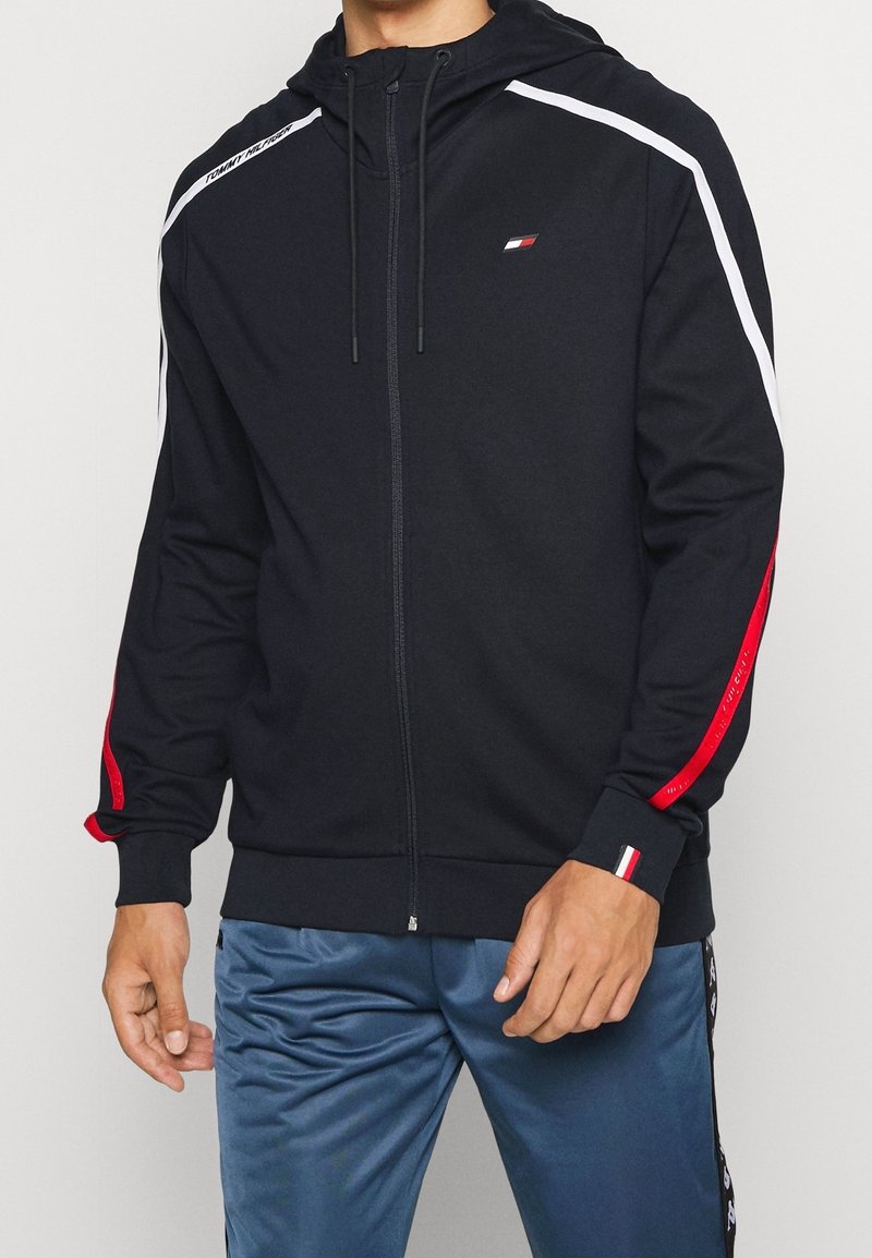 Schwarze Zip-Hoodie mit Kapuze, roten und weißen Akzentstreifen an den Ärmeln und einem kleinen Logo auf der Brust. Hergestellt aus weichem Stoff.
