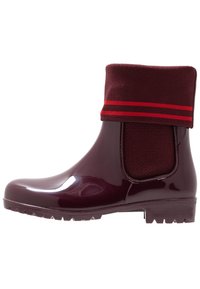 Botas de lluvia brillante en color burdeos hasta el tobillo con puño de punto que presenta dos franjas rojas horizontales.