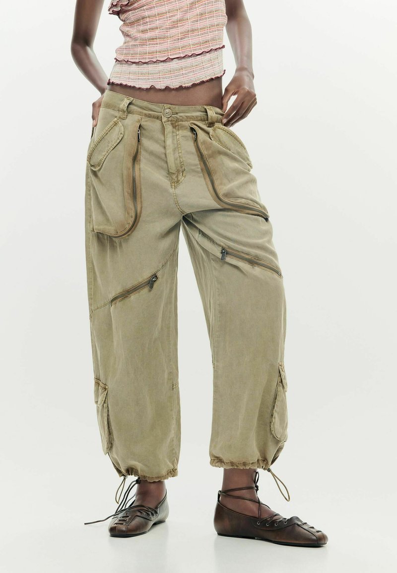 Persona vistiendo pantalones cargo sueltos de color verde oliva con múltiples cremalleras y puños con cordón, combinados con zapatos planos marrones con cordones y un top rosa corto.