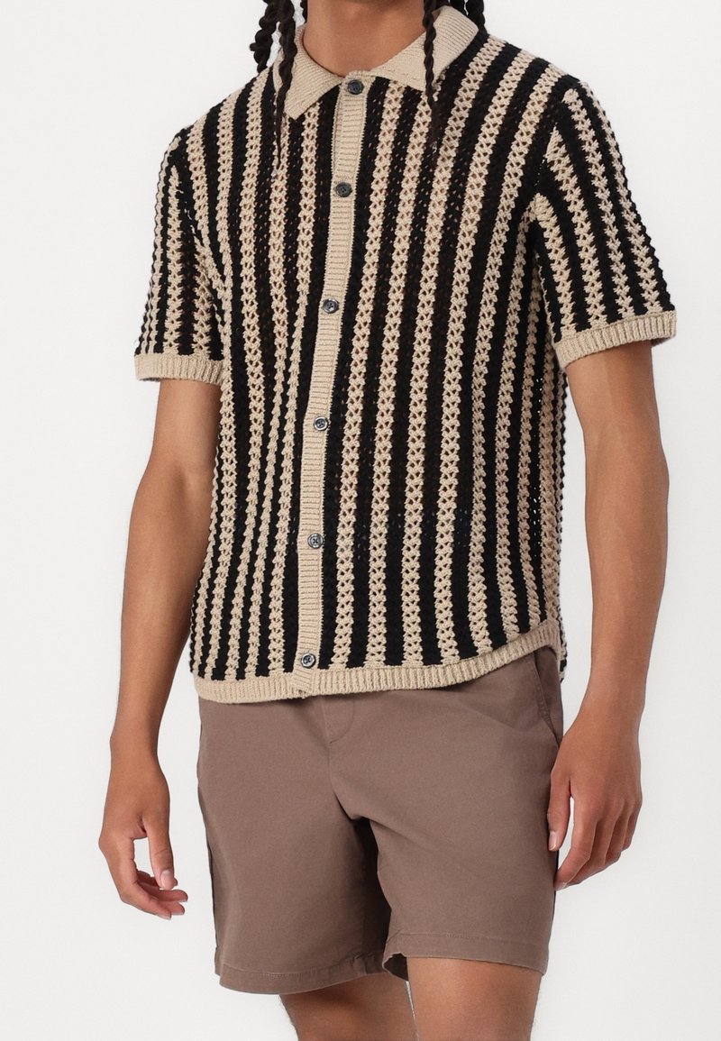 Gebreid shirt met korte mouwen in zwart en beige met een gestreept patroon, knoopsluiting aan de voorkant en geribbelde kraag en manchetten. Gecombineerd met bruine shorts.