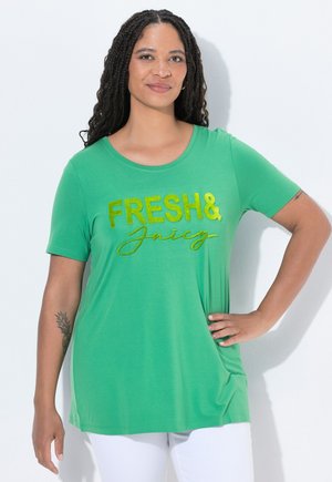 Vrouw met lang gevlochten haar, draagt een groen shirt met gouden tekst "FRESH & Juicy", witte broek en tatoeages op haar linkerarm, poseert met haar hand op haar heup.