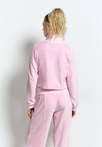 Kobieta z blond kręconymi włosami w kucyku, ubrana w jasnoróżowy, długi rękaw, krótką bluzkę i dopasowane szerokie spodnie, stojąca przed białą ścianą.