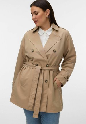 Femme portant un trench-coat beige à double boutonnage avec une ceinture nouée à la taille, une chemise blanche à col en dessous, et un jean bleu.