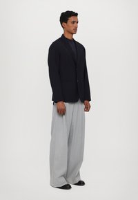 Zwarte blazer met twee knopen, zijzakken en geknipt revers; gecombineerd met lichtgrijze wijde broek en zwarte schoenen.