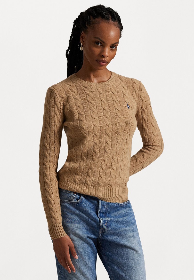 Pull en tricot beige avec un col rond, des poignets et un ourlet côtelés, orné d'un petit logo sur la poitrine. Associé à un jean bleu.